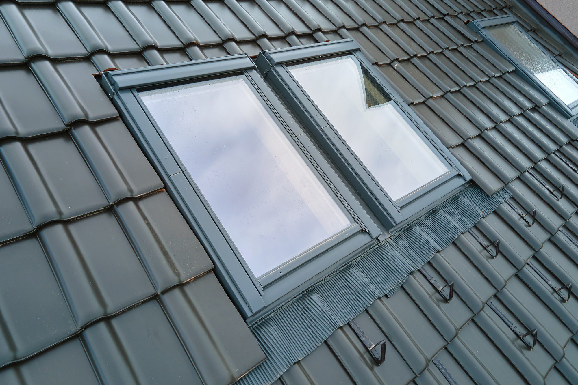 This image shows a close-up view of two windows installed on a pitched roof that is covered with grey metal tiles. The windows are designed as skylights, built to let natural light into the rooms below. The metal roofing has a pattern of ridges that provide structural support and a way for water to run off. There are some weatherproofing and sealing materials around the window frames ensuring they are watertight and insulating the interior against the elements. The sky is reflected in the glass of the windows, and there are some visible raindrops as well, suggesting recent or ongoing rain.