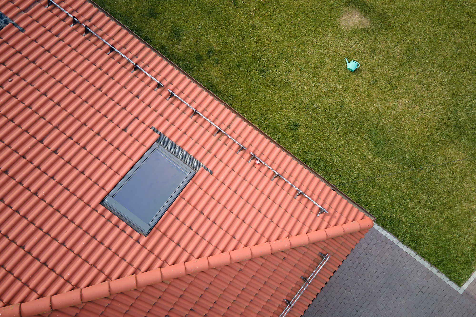 This image shows a bird's-eye view of a portion of a building and its surroundings. The top two-thirds of the image features a terracotta-coloured tiled roof with a single window or skylight. An array of metallic piping or guttering runs along the surface of the roof. Below the roof, there's a grassy area where the lawn is well-maintained, with one visible dry circular patch. On the right side of the grass area, there's a section of grey paving stones or patio. A blue watering can is lying on the grass, providing a splash of colour against the green. The perspective of the photo suggests it was taken from a higher point, looking down at the roof and the ground nearby.