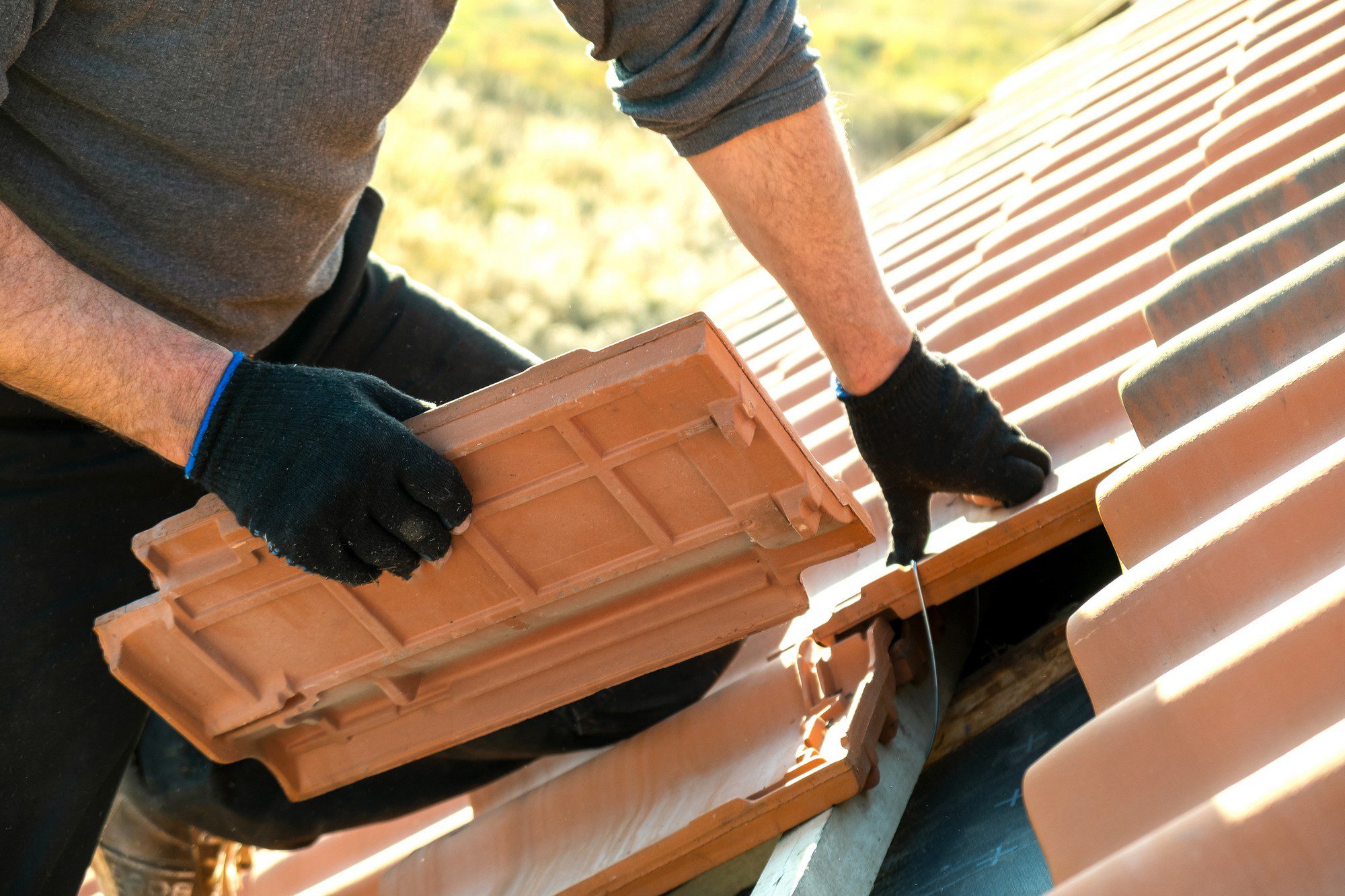 This image depicts a person engaged in roofing work, specifically installing or repairing clay tiles on a roof. The individual is wearing gloves for protection, and you can see the hands holding a terracotta-coloured roof tile, which is about to be placed alongside other similar tiles in a neat arrangement typical of a tiled roof. The background suggests an outdoor setting, likely with greenery, although it is out of focus. The focus is on the hands and the tile, emphasizing the roofing work in progress.