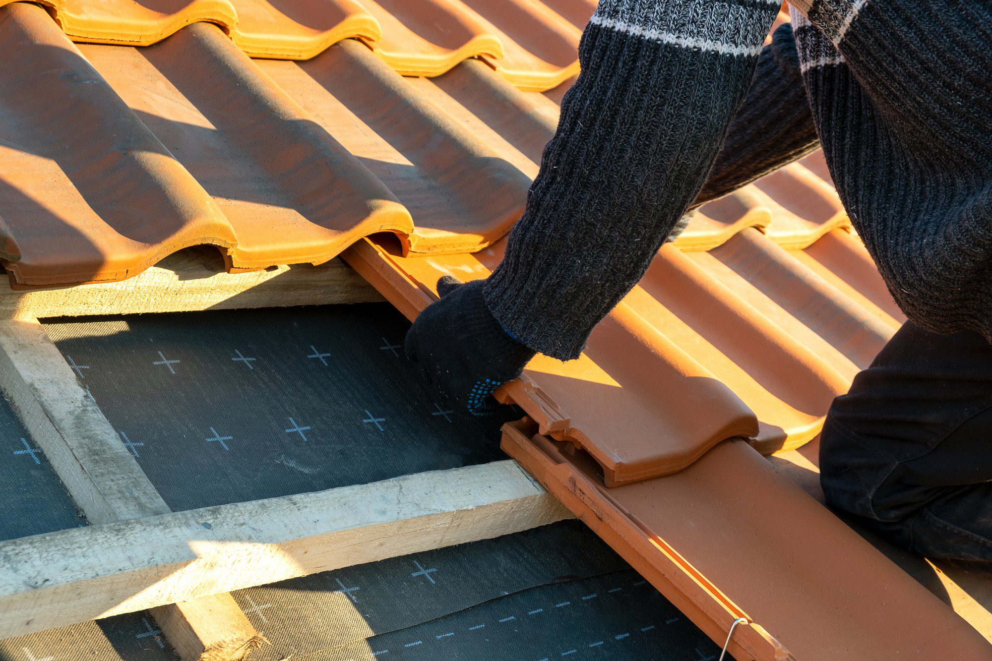 This image shows a close-up of a person working on a tiled roof. The person is wearing a knitted jumper and gloves, suggesting that the weather may be cool. They are in the process of either repairing or constructing the roof, as indicated by the exposed underlayment and open section where roof tiles have not yet been placed.The roof tiles are orange-coloured and have a wavy pattern typical of clay or concrete tiles, commonly used for their durability and aesthetic appeal. Wooden battens are installed to provide a fixing point for the tiles. The section of the underlayment that is visible is marked with "+" signs, which might be for alignment or measurement purposes.The worker appears to be handling one of the tiles, suggesting active work happening on the roof. The photograph captures a moment of manual labour in the construction and maintenance of housing infrastructure.