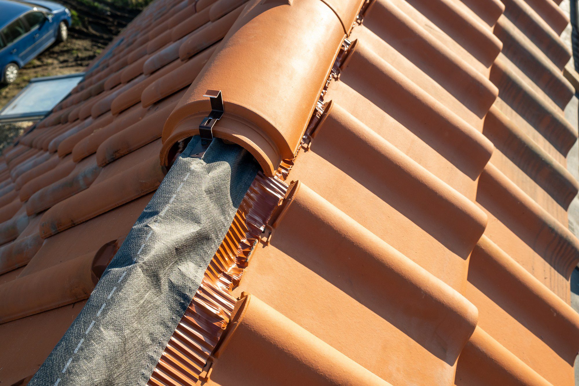 This is an image of a ceramic tiled roof. You can see the rounded tiles aligned neatly side by side, and a ridge vent covered by a protective fabric to prevent debris from entering while allowing air to flow. The structure underneath the vent is likely to be a ridge or hip on the roof where two slopes meet. The photo is taken from a high angle looking down the slope of the roof, showcasing the arrangement and construction of the tiles and the ridge vent. There's also a view of a car parked below in the background, providing some context about the height from which the photo was taken.