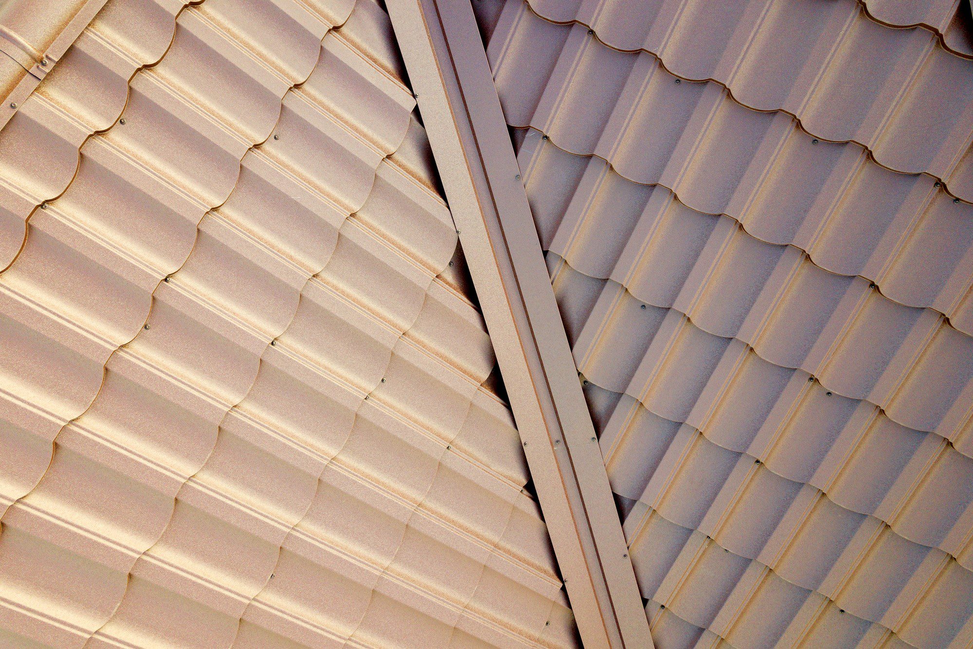 The image shows a close-up of a corrugated metal roof. There are two distinct sections with a metal beam or flashing separating them, suggesting a change in slope or an intersection of two roofing sections. The metal has a beige or tan colour, and you can see the individual panels joined together by screws or rivets at regular intervals along their ridges. The pattern and repetition create an interesting geometric texture.