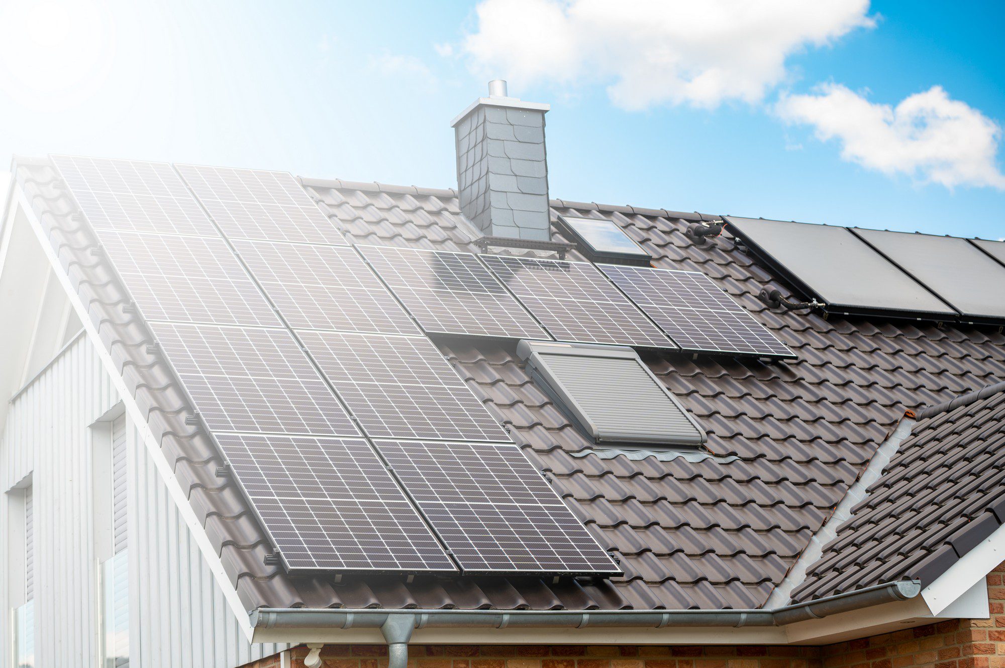 The image shows the roof of a residential building equipped with solar panels. These panels are installed to capture sunlight and convert it into electricity, as part of a solar energy system for the home. It's a sunny day and the sky is partly cloudy, suggesting good conditions for solar energy production. Additionally, there are some roof windows or skylights integrated into the roof, which are used for natural lighting inside the building. The chimney visible on the roof suggests that there might also be a traditional heating system installed in the house.