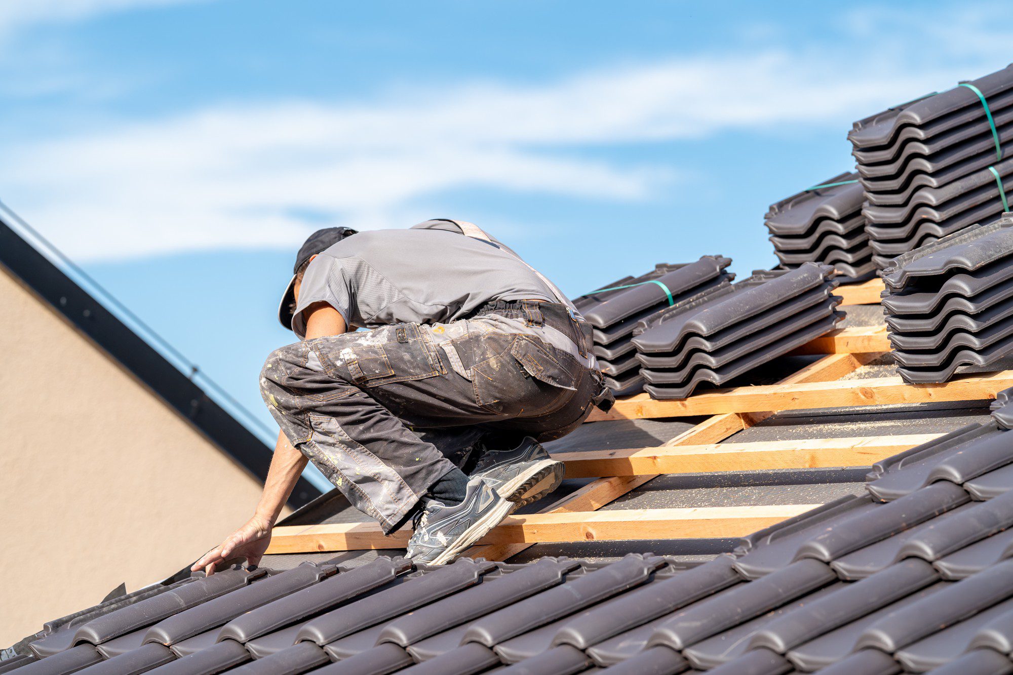 In the image, there is an individual working on a roof. The person appears to be installing or repairing roof tiles. They are bent over, focusing on their work. The roof tiles are dark in colour, which contrasts against the clear blue sky in the background. There are stacks of additional roof tiles on the structure, indicating that the roofing work is in progress. The person is wearing casual work attire including a cap, possibly for protection from the sun. Safety measures such as the use of a harness or other protective equipment are not visible in the image. The overall scene is one of manual labour, specifically roofing construction or maintenance.