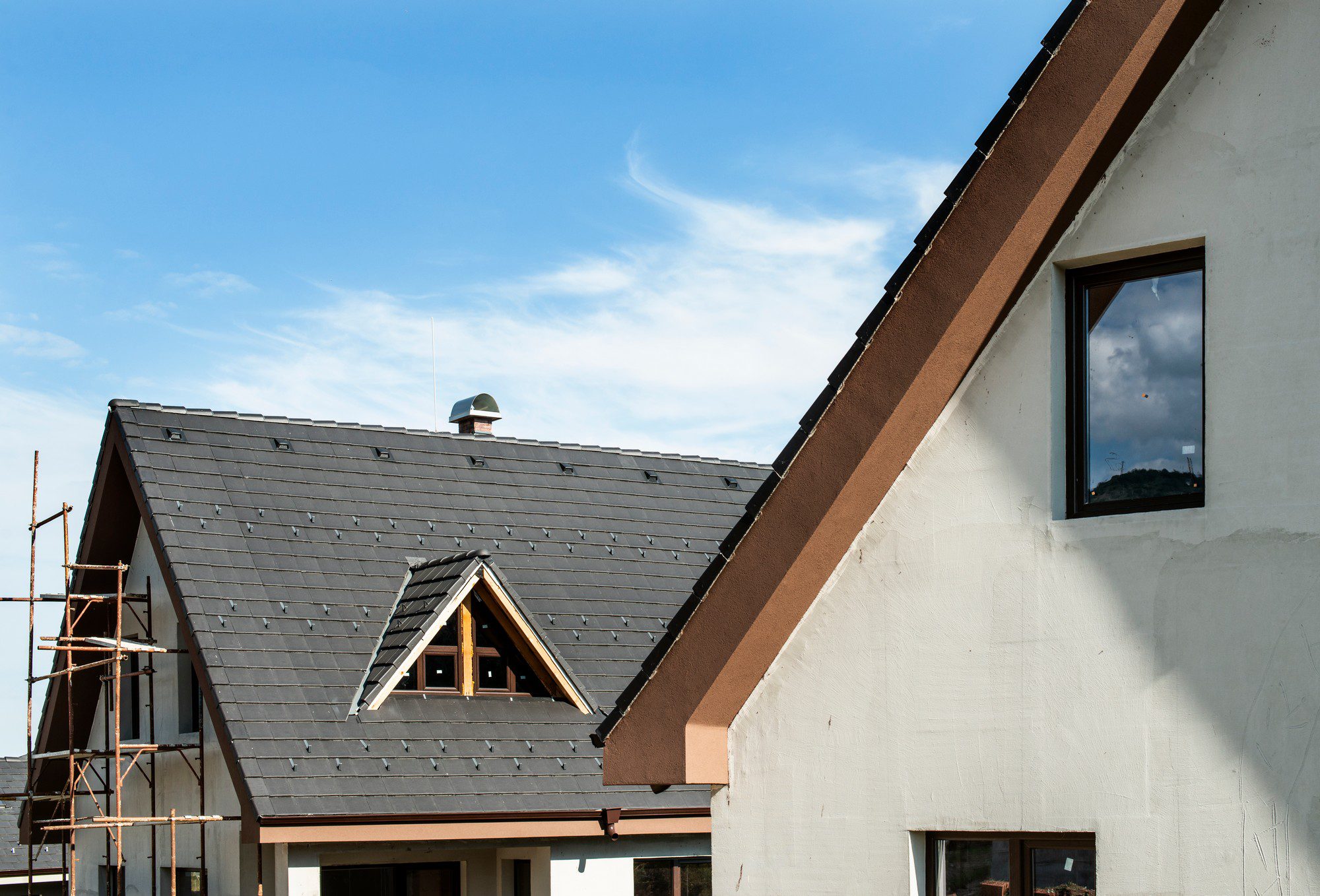 This image shows a part of a residential building under construction or renovation. There is scaffolding along the side of the building, which indicates work is being done on the exterior. The house has a pitched roof with a dark-coloured roofing material, and there is a roof window (dormer window) as well as a ventilation chimney visible on the roof. The side of the house displays a large window under the gable, and the exterior walls are finished with what appears to be a white render or plaster. The sky is mostly clear with some wispy clouds, suggesting it may be a fair weather day for construction.