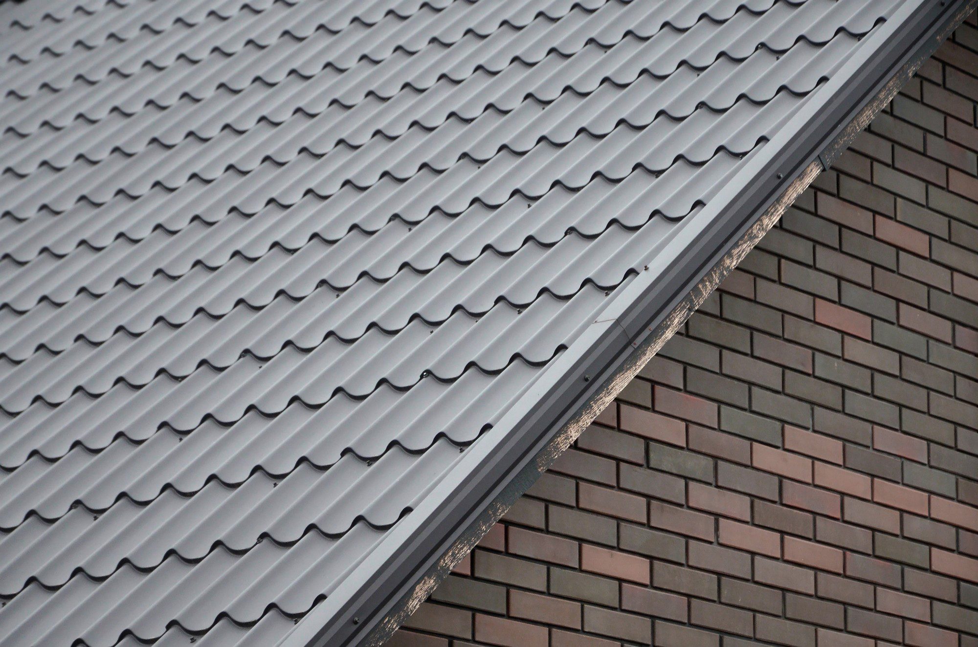 This image shows a close-up of a building's exterior materials. On the top half of the image, there is a section of a pitched roof covered with gray metal roofing tiles, characterized by their wavy, corrugated pattern. On the bottom half, there is a wall surface covered with brick cladding, featuring bricks in varying shades of browns and reds laid in a standard pattern. The intersection where the roof meets the wall is sealed and reinforced with a strip of metal flashing to prevent water infiltration. The elements in the image suggest a focus on building construction and roofing techniques.