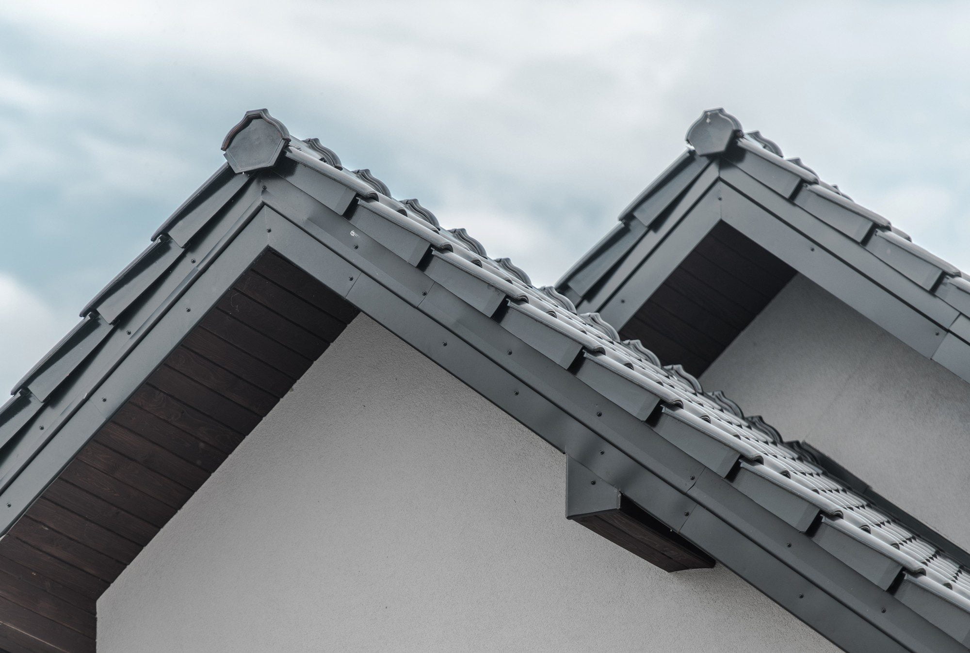 The image displays the upper portion of a building's facade featuring architectural details of its roof. There are two sloping sides of the roofline visible, both equipped with grey roofing tiles and capped with a ridge piece at the top where the two slopes meet. The eaves of the roof extend beyond the wall, and there are visible guttering and downspouts, essential for directing rainwater away from the building. The building's attic areas, just beneath the roof, have a wooden finish, providing a contrasting texture and colour to the lighter stucco walls. The sky in the background is overcast, suggesting cloudy weather.