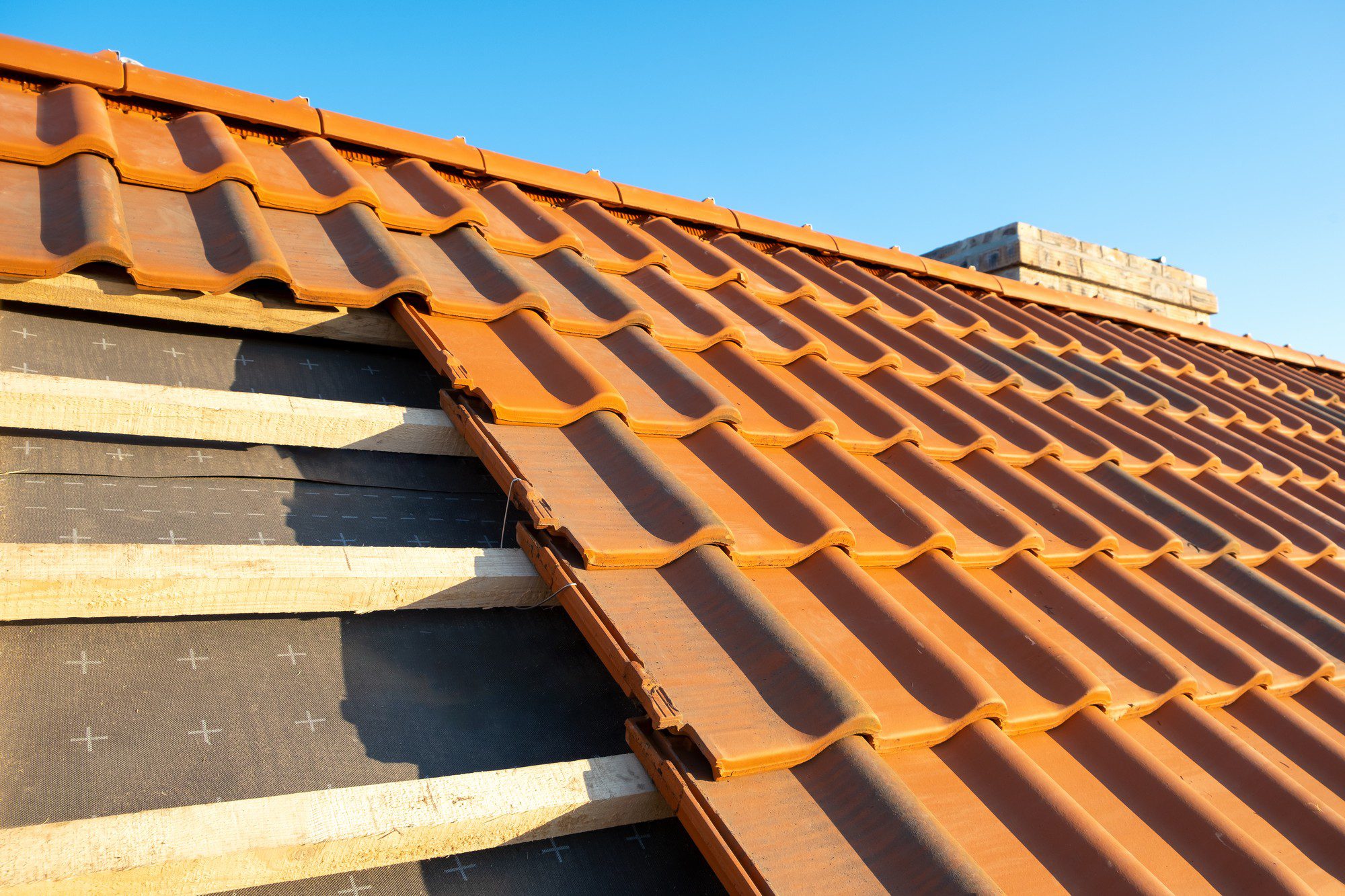 This image features a close-up view of a terracotta tile roof. You can see a series of curved tiles installed on the roof, and part of the roof's underlayment is visible where the tiles have yet to be laid. The underlayment is black and seems to be marked with a grid pattern that might be used for aligning the tiles. The image is taken against a backdrop of a clear blue sky, indicating good weather conditions, which is often preferred for roofing work. The angle of the image provides a perspective looking up the slope of the roof.