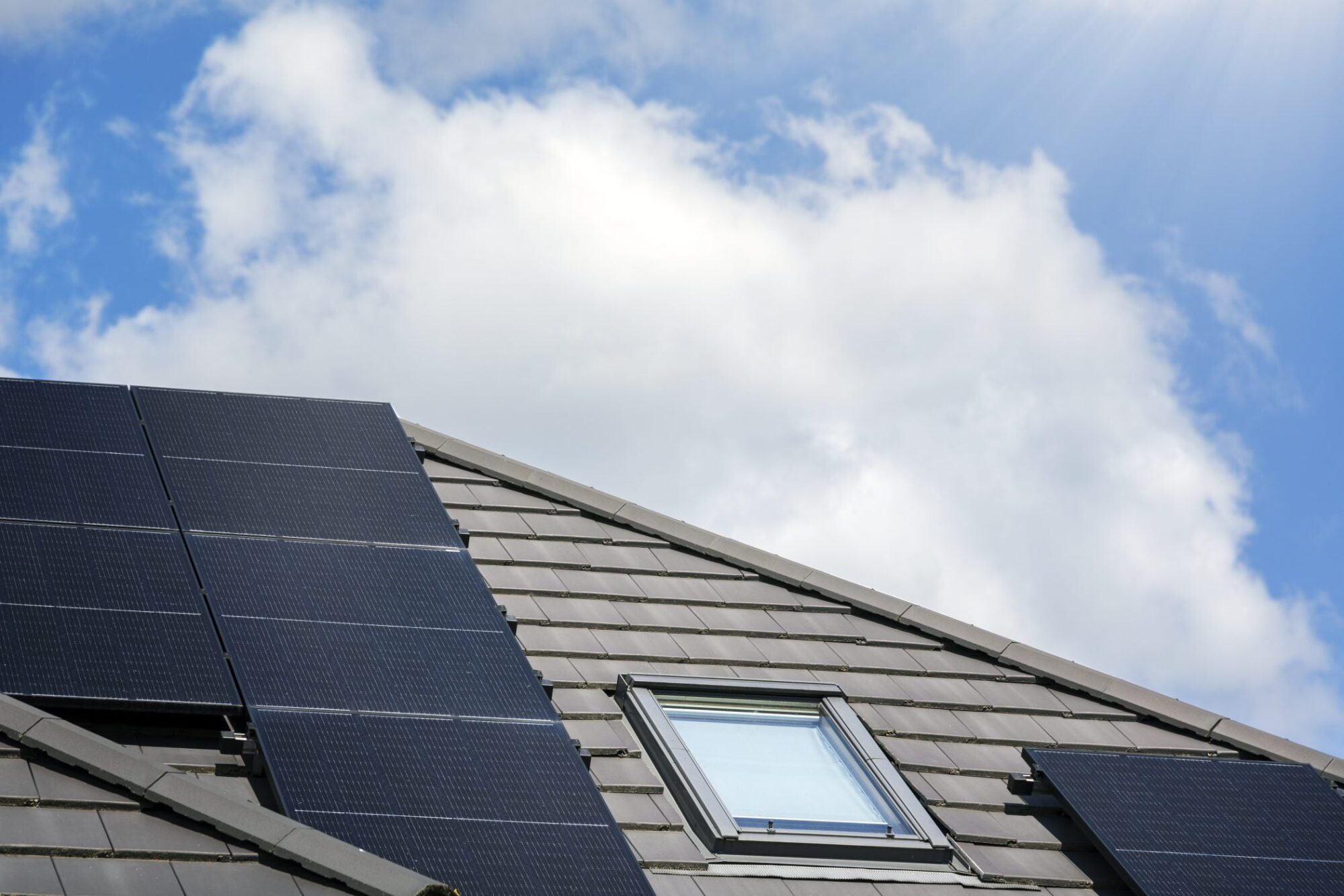 The image shows a section of a sloping roof equipped with solar panels. There is a skylight window incorporated into the roof, allowing natural light to enter the space below. The background depicts a blue sky with some scattered clouds and the sun shining from the upper right corner, suggesting a bright day which is ideal for solar power generation. The solar panels are installed on top of the roof tiles and are positioned to maximise exposure to the sunlight.