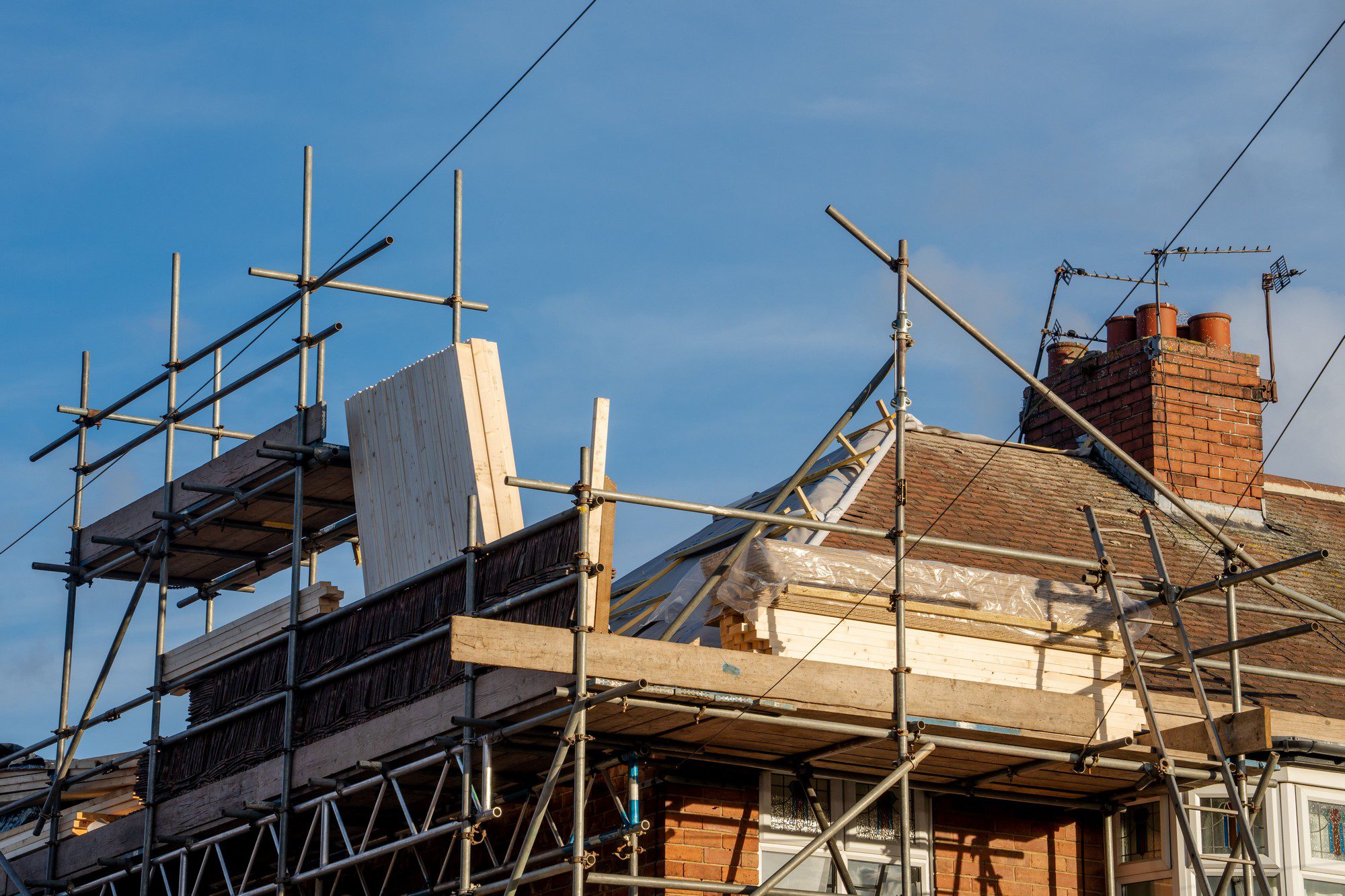 This image shows the exterior of a house currently under construction or renovation. There is scaffolding set up around the upper part of the building, indicating work being done on the structure, likely on the roof or walls. The scaffolding is made up of metal poles and wooden planks to provide a platform for the construction work. We can also observe that the house has a brick chimney with multiple flues, as well as a television antenna on the roof. The sky is clear indicating fair weather which is usually suitable for construction activities.