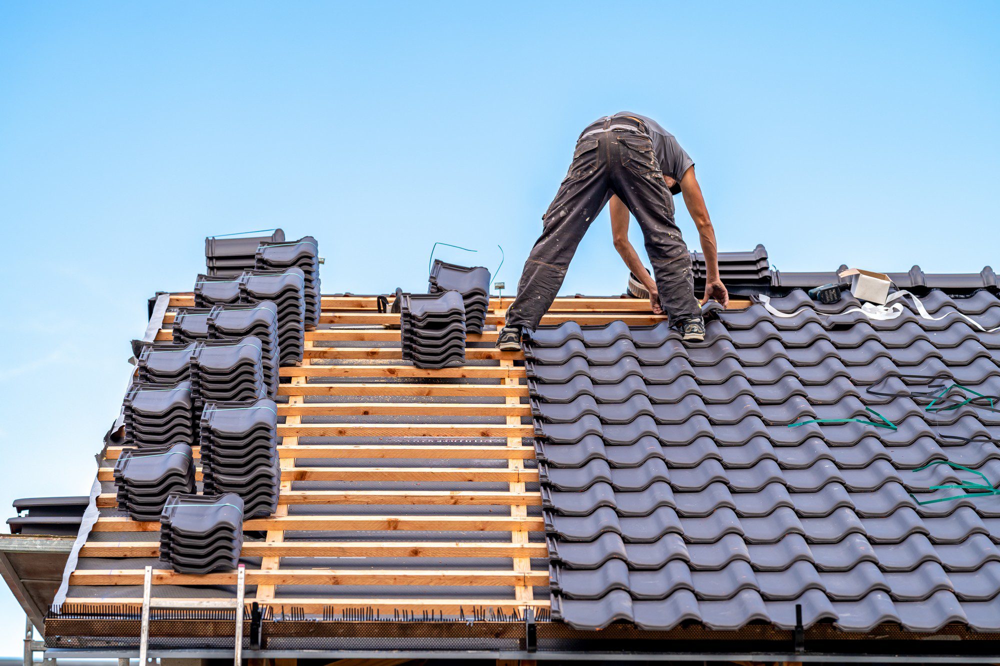 The image shows a person working on a roof installation or repair. They seem to be installing a new layer of roofing tiles. The roofing tiles appear to be of the interlocking type, commonly used due to their durability and resistance to weather. The individual is bent over, carefully placing a tile while standing on the wooden roof battens where the tiles are being laid. The roof structure and the way the tiles are stacked suggest that work is in progress. The background is a clear blue sky, indicating good weather conditions for outdoor construction work. Safety precautions, such as proper footing and perhaps safety gear, are important for the individual working at such heights.