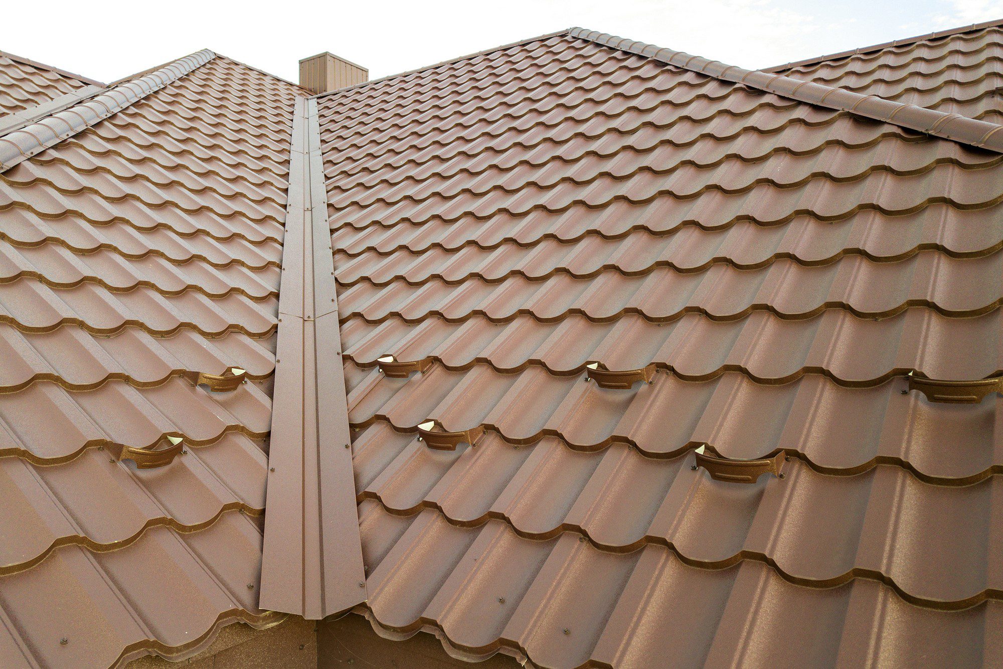 The image shows a close-up view of a section of a building's roof. The roof is made of terracotta-coloured tile with a wavy, S-shaped profile, which is typical of clay or concrete roofing tiles. There are several rows of tiles visible, and you can see ridge caps where two sloped sections of the roof meet, indicating this is likely the peak of the roof. Additionally, there are snow guards installed on the tiles: these are devices intended to prevent snow slides by allowing the snow to melt gradually or break apart in small amounts rather than falling off the roof in large sheets, which can be dangerous and cause damage. The sky suggests either early morning or late afternoon, and the light colour of the tiles might indicate that they have been coated for reflective purposes or just the colour choice for aesthetic reasons.