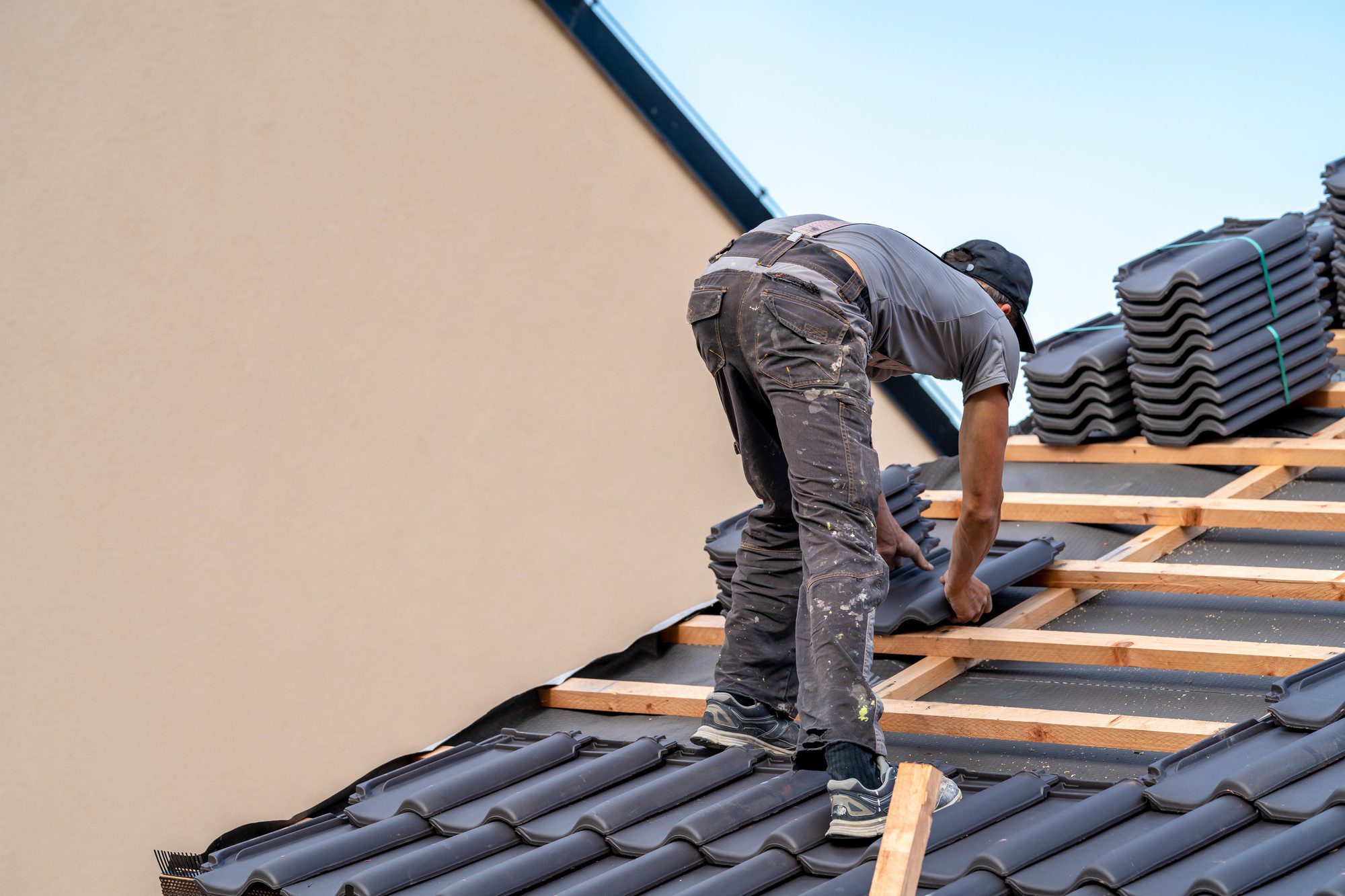 The image shows a person installing roof tiles. The individual is bent over, working on a sloped roofing structure made of wooden battens. They are wearing a cap, and protective clothing covered with dust or paint spots, hinting at their hands-on work. A stack of additional roof tiles is piled neatly on the roof to the right. It appears to be a bright day, and the safety measures for such work are not directly visible in the photo. The person is likely a skilled worker or roofer, and the setting suggests construction or roofing maintenance activities.