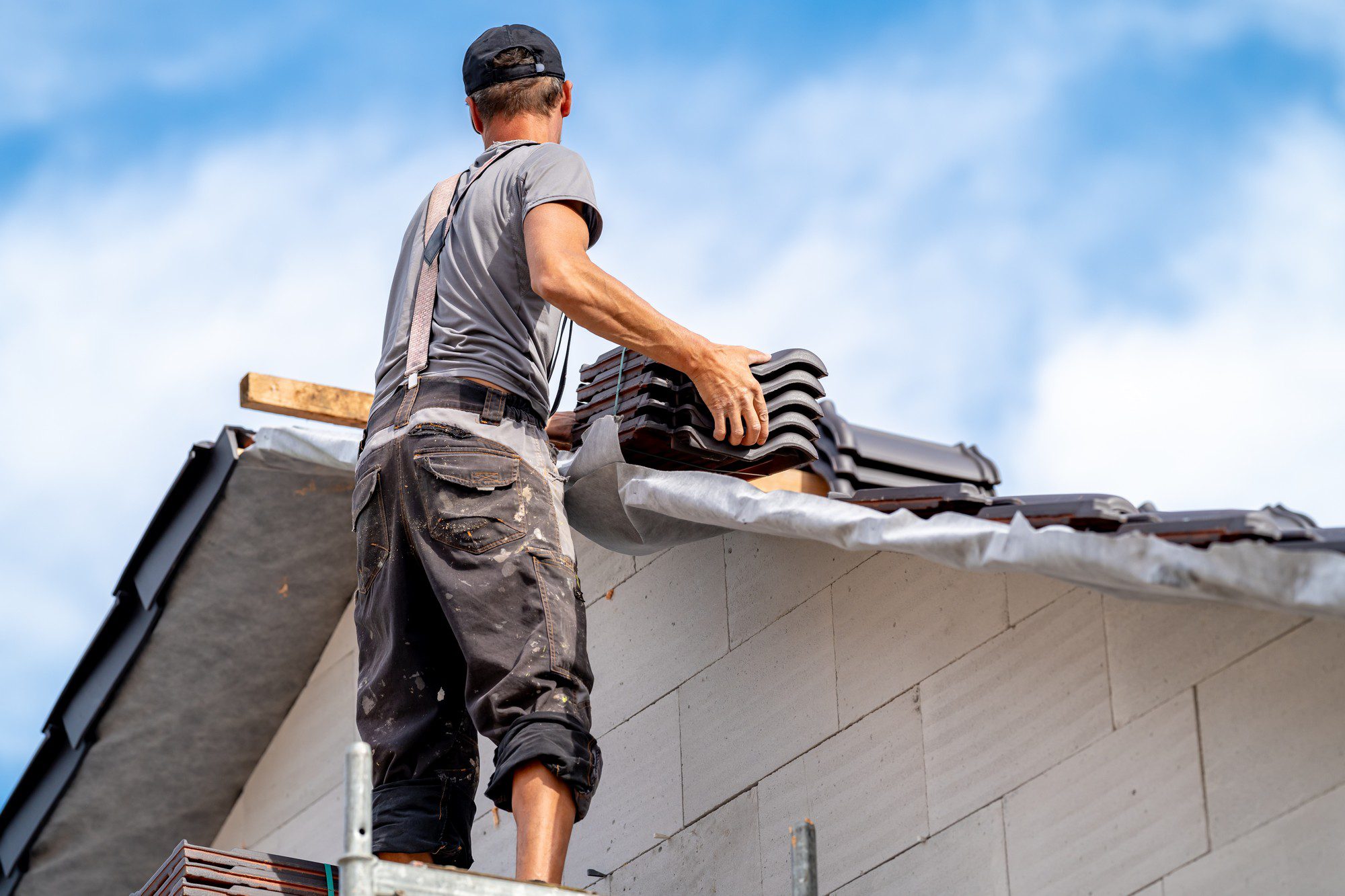 The image shows a person working on a roof. The individual appears to be installing or repairing a tiled roofing system. They are wearing a grey shirt, work pants with various pockets likely for tools, a dark cap, and a pair of braces. The person is handling roofing tiles and seems to be laying them on the roof structure. Below the person, we see a portion of the roof underlayment or roofing felt, which is used to provide a secondary waterproof barrier. The background includes a clear blue sky with wisps of clouds. The focus is on the worker's activities, illustrating labour common in construction and roofing projects. The person's face is not visible as they are turned away from the camera, focusing on the task at hand.