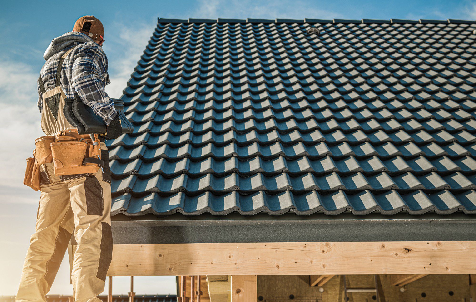 The image shows a person standing on a scaffold and working on a house roof. The person is dressed in a plaid shirt, has a jacket tied around the waist, and is wearing a tool belt loaded with various tools. They also wear protective gear such as a hard hat. The roof is covered with dark-coloured, curved tiles, commonly known as Spanish tiles or barrel tiles. The sky is clear with few clouds, suggesting good weather for outdoor construction work.