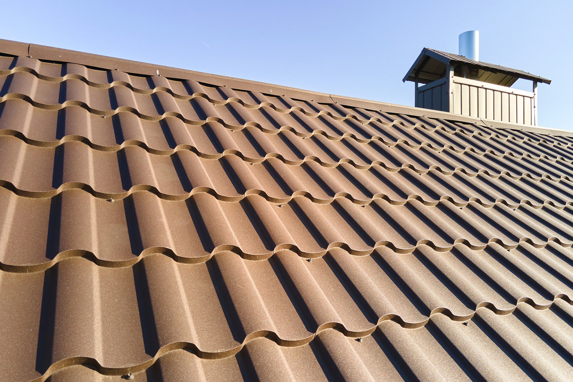 The image shows a close-up view of a brown tiled roof with a clear blue sky in the background. The tiles have a corrugated pattern, which is typical for terracotta or concrete tiles, designed to interlock and channel water effectively. On the right side of the image, there is a chimney poking out above the roof, suggesting that the building likely has a fireplace or some sort of exhaust that requires venting through the roof. The roof appears to be in good condition, without any visible damage or wear.