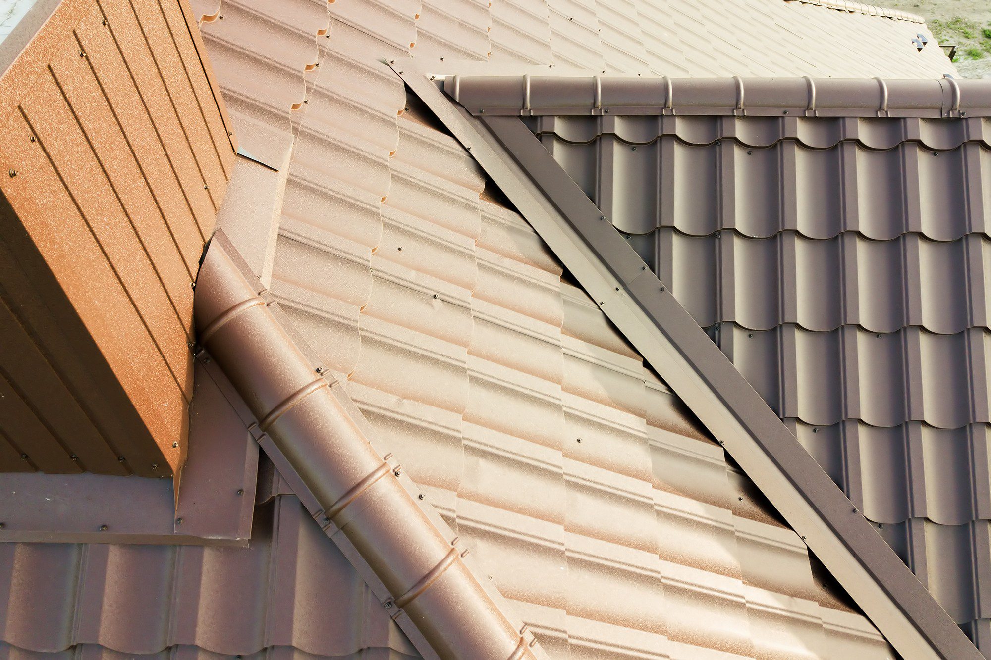 This image shows a close-up view of a building's roof. The roof is made of metal tiles, which are commonly used for their durability and longevity. There are two distinct colours and patterns of metal roofing visible, implying that either an extension was added or a repair was done using different materials. A metal gutter system is also in place to channel rainwater away from the structure, with downspouts for proper drainage. The construction appears to be meticulous, with all the tiles and gutter components neatly aligned and secured.