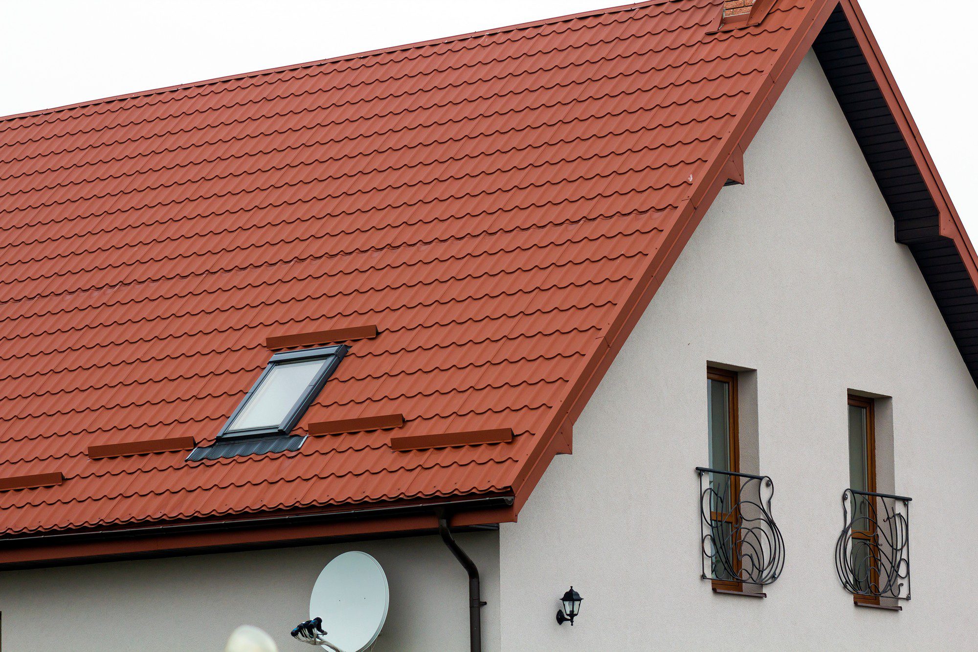 The image shows the upper part of a building featuring a steeply pitched roof with terracotta-coloured tiles. There is a roof window (skylight) with a black frame on the left side of the roof. The eaves are equipped with snow guards, which are metal bars installed to prevent snow from sliding off the roof in large quantities. The building's facade is a light colour, and there are two windows each with an ornamental metal balcony railing. To the left, mounted on an exterior wall, there is a satellite dish, and underneath it, a black outdoor wall lantern is visible, providing exterior lighting.