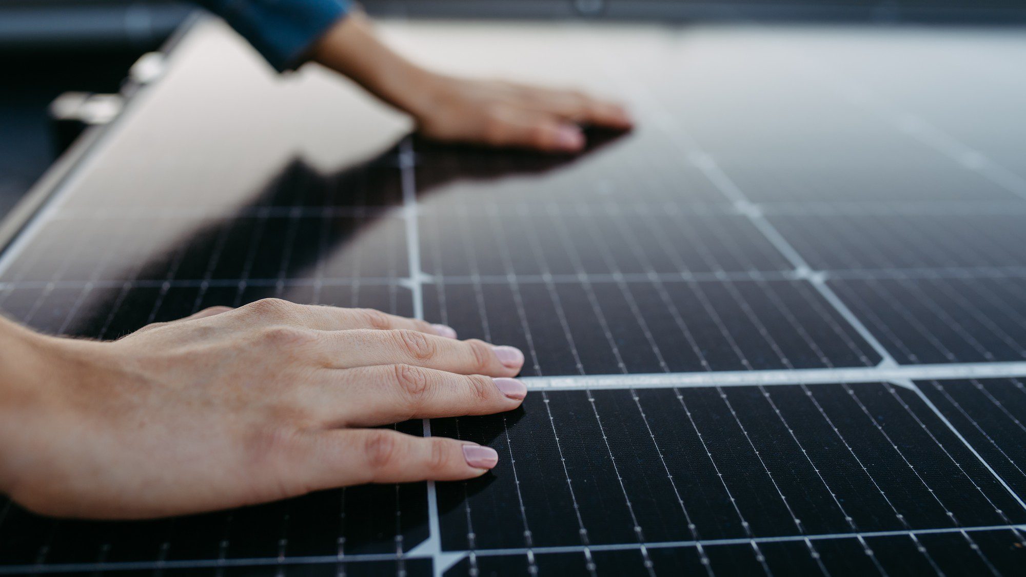 This image shows a pair of human hands placed gently on a surface of solar panels. The solar panels have a distinct pattern of cells and are typically blue-black in colour, which is common for silicon-based photovoltaic cells. The person's hands touching the solar panels could suggest an inspection, installation, maintenance, or a connection gesture to renewable energy technology.