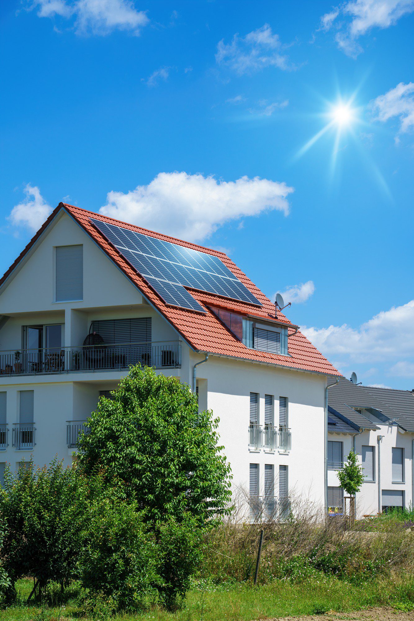 This image features a modern two-story residential building with a pitched roof that is equipped with solar panels. The house has multiple windows, some with shutters, and what appears to be a balcony on the upper floor. A lush green tree and some shrubbery are visible in front of the house, indicating a well-vegetated area. In the background, the sky is clear and blue with few clouds, and the bright sun is shining, which is ideal for generating solar energy. The environment suggests a suburban or residential setting. Overall, the image portrays a theme of sustainable living and renewable energy use in a domestic setting.