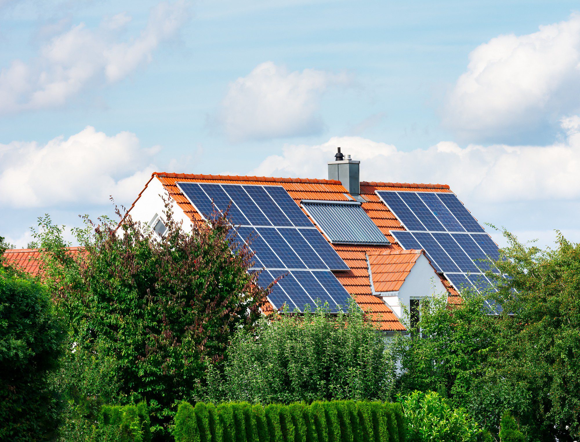 The image displays a residential house with a pitched roof that is equipped with multiple solar panels. These panels are installed on the roof to absorb sunlight and convert it into electricity, reflecting a move towards sustainable energy use. The house appears to be in a suburban setting, with a variety of lush green plants and trees surrounding the property, which could indicate the homeowner's preference for green living. A vibrant blue sky with scattered clouds can be seen in the background, providing a pleasant and serene atmosphere.