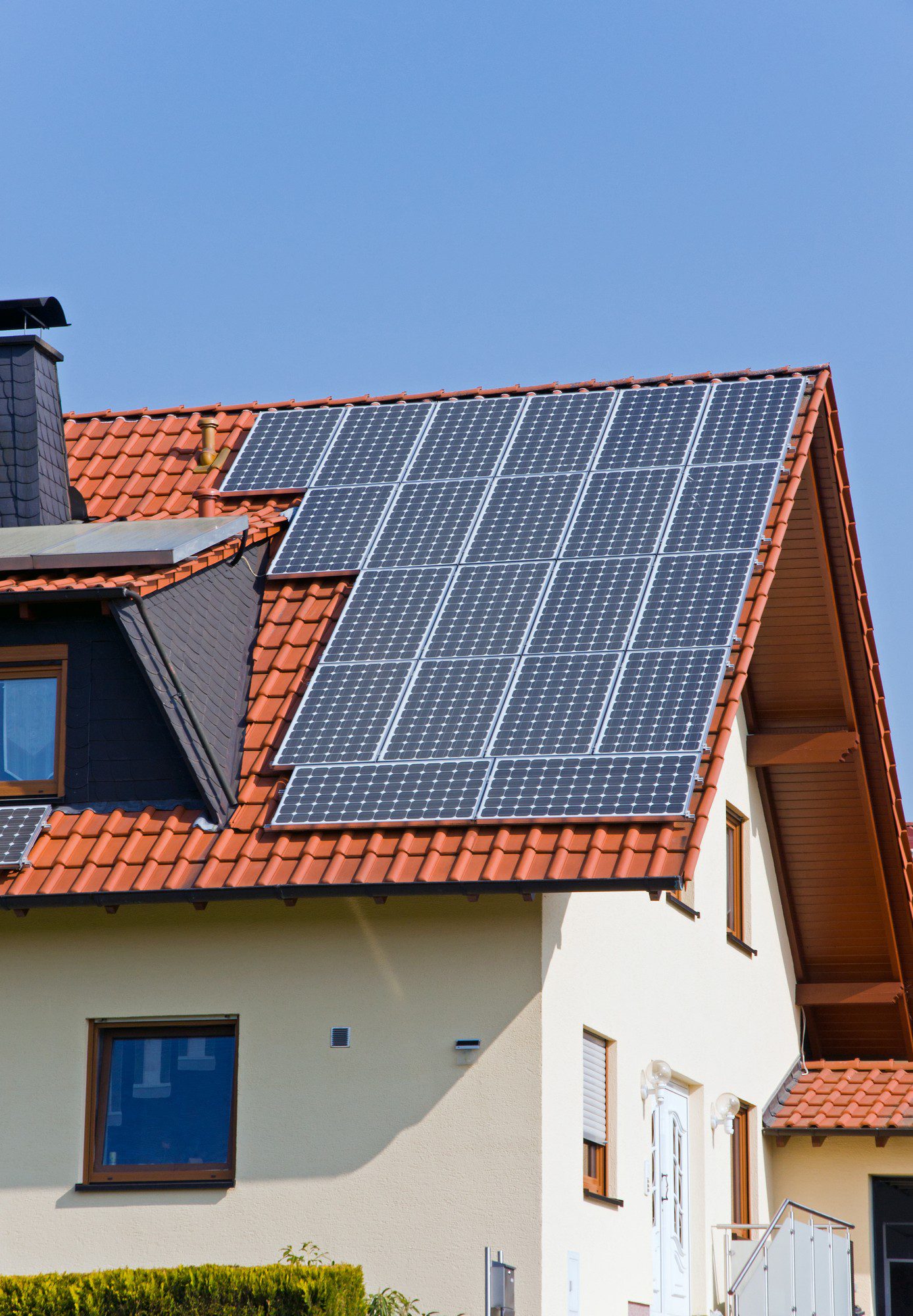 The image shows a section of a residential building with solar panels installed on the slope of its roof. The solar panels are designed to capture sunlight and convert it into electricity. The roof is covered with terracotta-coloured tiles, and there is a clear blue sky in the background. The house appears to be a detached single-family home with a visible window, beige exterior walls, and a partial view of an entrance with steps and a handrail. The solar panels indicate an investment in renewable energy and sustainability.