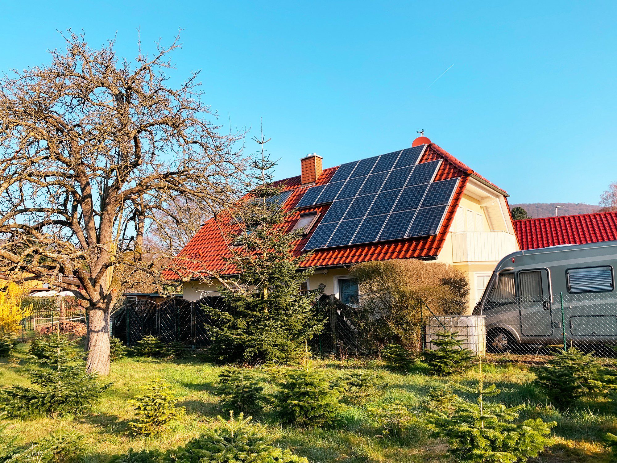 This image shows a residential setting with a house that has a red-tiled roof and equipped with solar panels. There's a leafless deciduous tree near the house, suggesting it might be late autumn or winter when the photo was taken. There is also a variety of greenery including coniferous trees and shrubs. On the right side of the photo, there's a parked caravan with a gray exterior. The weather appears to be sunny, and the sky is clear blue.