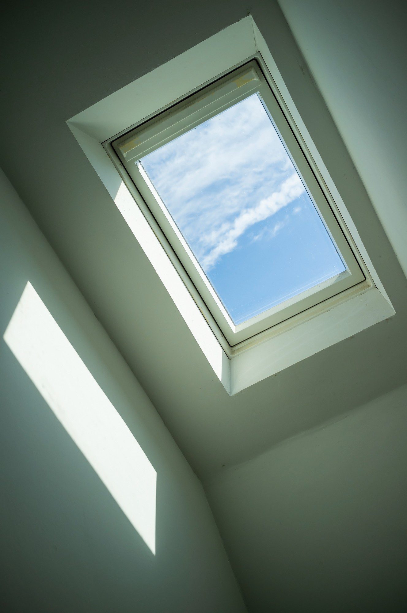 This image features a skylight window installed in a sloped ceiling. The window is providing a view of the sky and some clouds, suggesting it's a nice day outside. Light is entering through the skylight, casting a sharp, angular shadow on the white ceiling beneath. The walls also appear to be painted white, which enhances the brightness of the space. The overall feeling conveyed by the image is of a calm and serene indoor area with natural lighting.