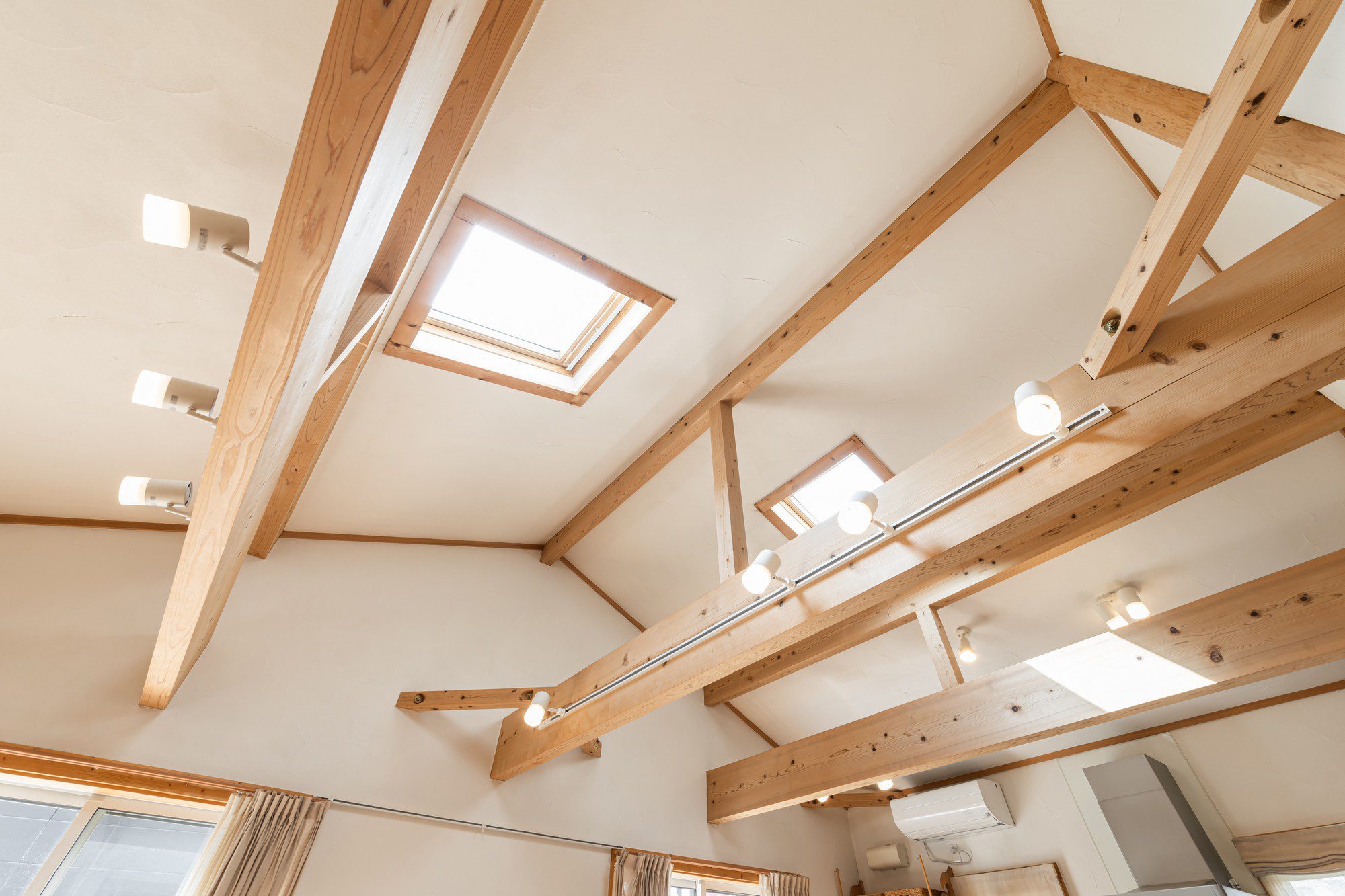 The image shows the interior ceiling of a room with a sloped, high ceiling. It features exposed wooden beams that provide structural support and an aesthetic quality to the space. There are track lighting systems mounted on some of these beams, equipped with multiple adjustable lights. Additionally, there are two skylights installed in the ceiling, allowing natural light to enter the room. The colours are warm, with the natural colour of wood contrasting with the off-white of the ceiling. The lower portion of the image shows curtains, hinting at windows or glass doors, and portions of a room that is not fully visible, possibly including furniture like cabinets.