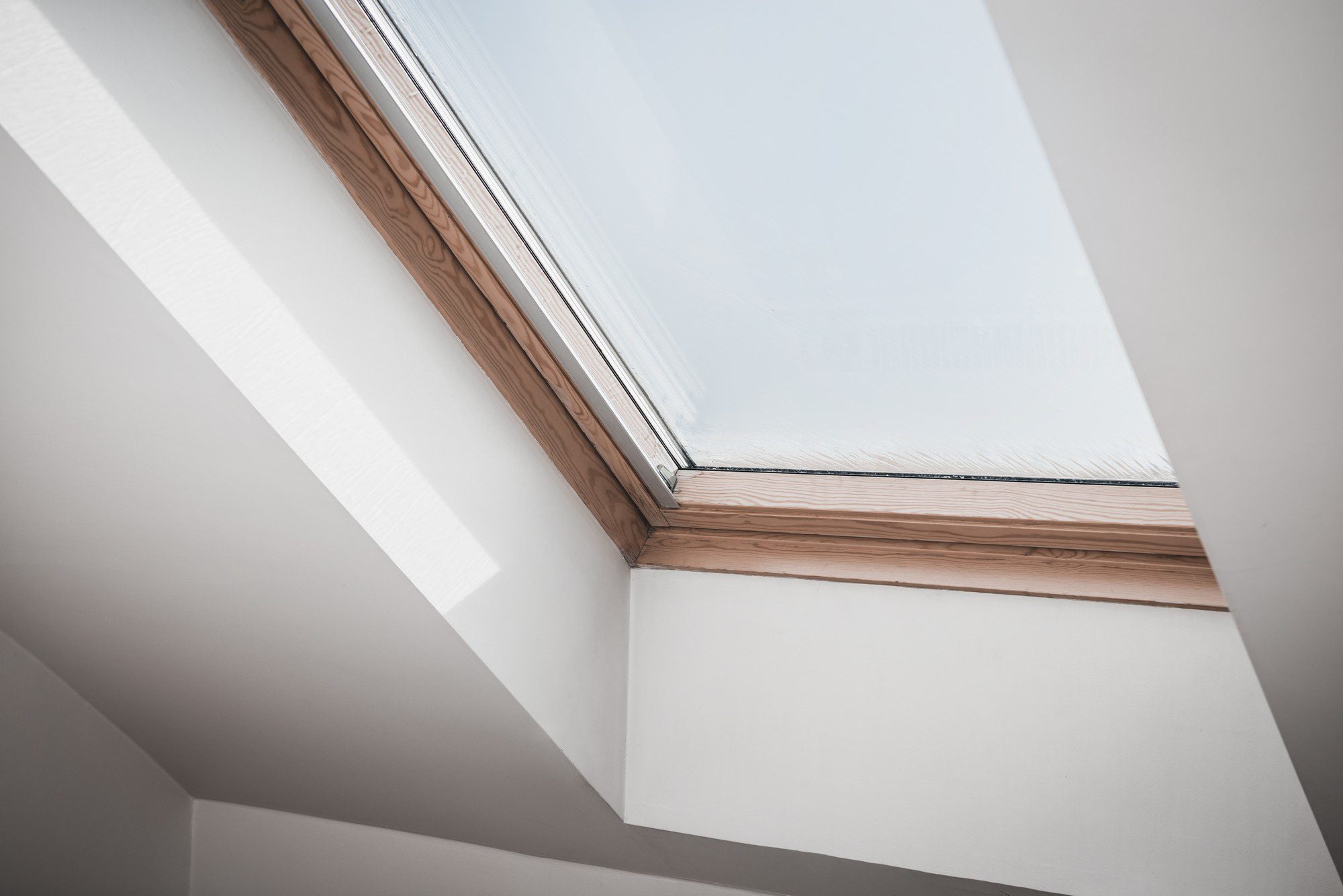 The image shows a modern skylight window installed in a sloped ceiling. The window has a wooden frame and appears to be closed. It is set into a wall with a white finish, and the angle of the photograph creates a geometric composition with lines and shapes formed by the intersection of the walls and the window. Sunlight is entering through the window, casting a bright rectangle of natural light onto the interior surfaces, accentuating the clean, minimalistic design. This type of window is commonly used to bring natural light into attic spaces or rooms with sloped roofs.