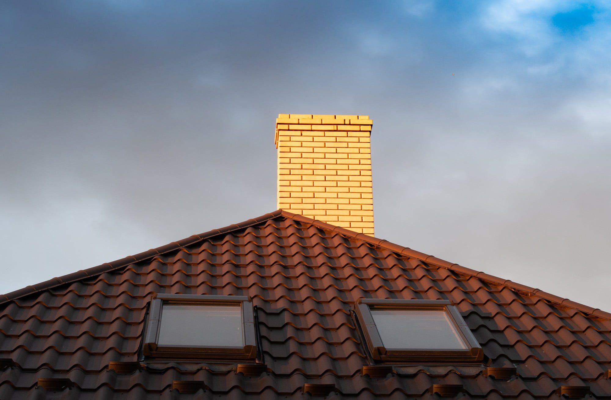 The image shows a portion of a building's roof with terracotta tiles. There are two windows, likely skylights, integrated into the roofing. Above the roof is a brick chimney that stands out against the blue and gray sky in the background. The warm light suggesting either sunrise or sunset casts a glow on the chimney, enhancing its colour contrast with the roof and sky.