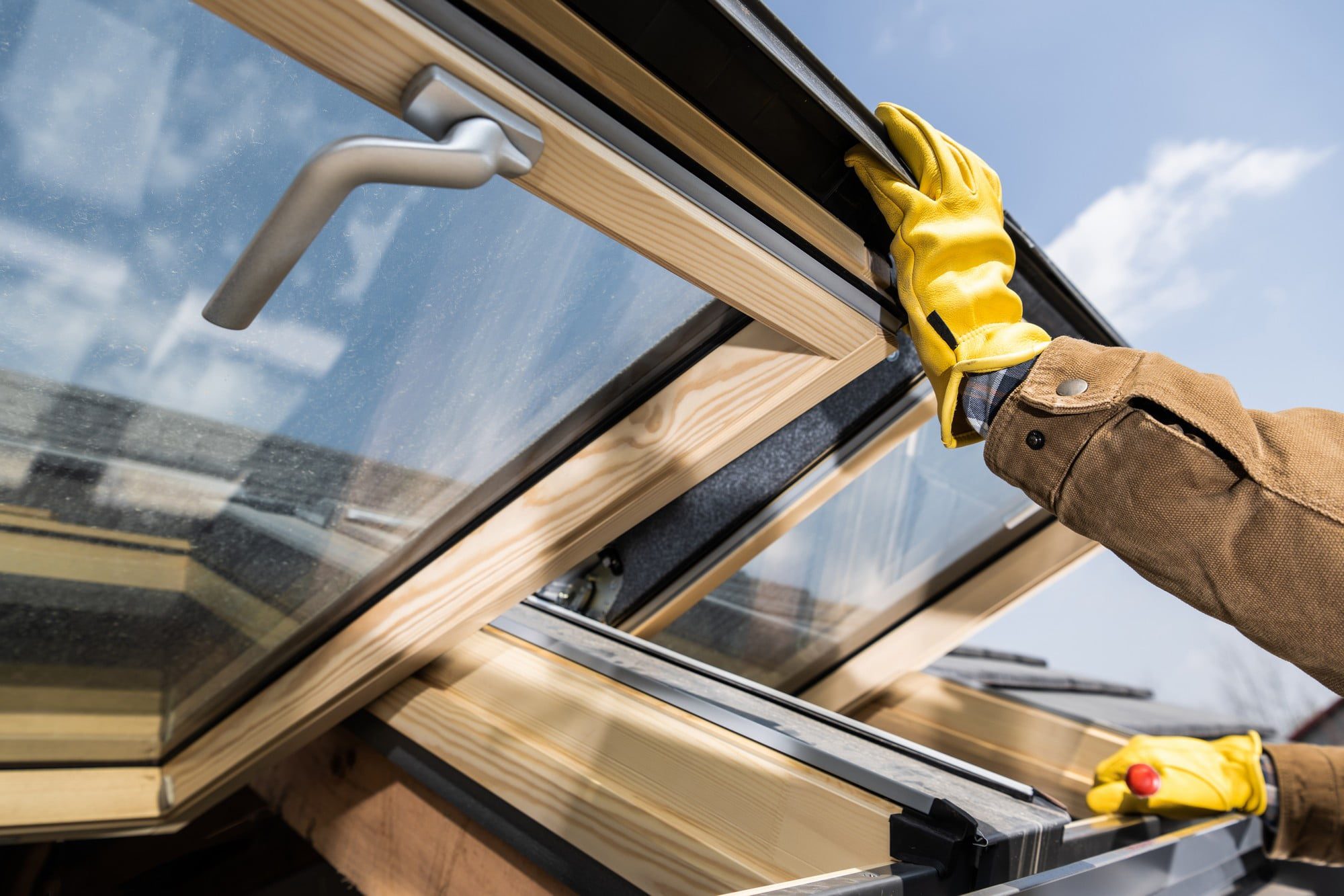 The image shows a person wearing yellow gloves and a brown long sleeve shirt working on a window. The person is likely engaged in installation, repair, or maintenance work. The window appears to have a wooden frame and a handle, indicating that it may be operable, probably a type that can be tilted or opened. The person is handling the window from the outside, with a clear sky visible in the background, suggesting the work is being done on an upper level or a roof window. The gloves and attire suggest the importance of safety and protection in the work being carried out.