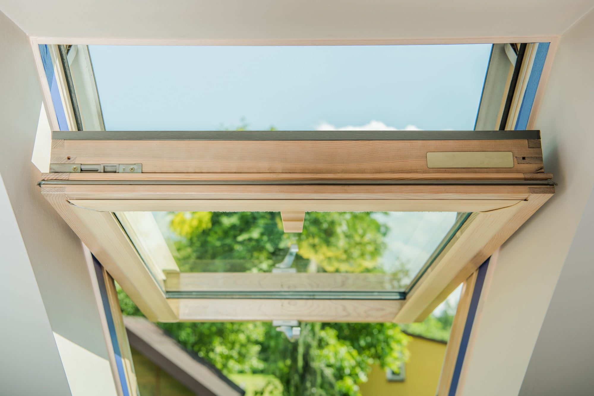 The image shows a partially open skylight window installed in a sloped ceiling. The window frame appears to be made of wood, and you can see a metal window hinge mechanism that allows the window to be held open at an angle. Through the glass of the skylight, a clear blue sky and the tops of green trees can be seen, suggesting a pleasant day outside. The interior around the window is finished with a white trim, giving a clean and modern appearance to the space. The scene suggests a room in an upper level of a house, such as an attic or loft, where skylight windows are commonly used to bring in natural light and ventilation.