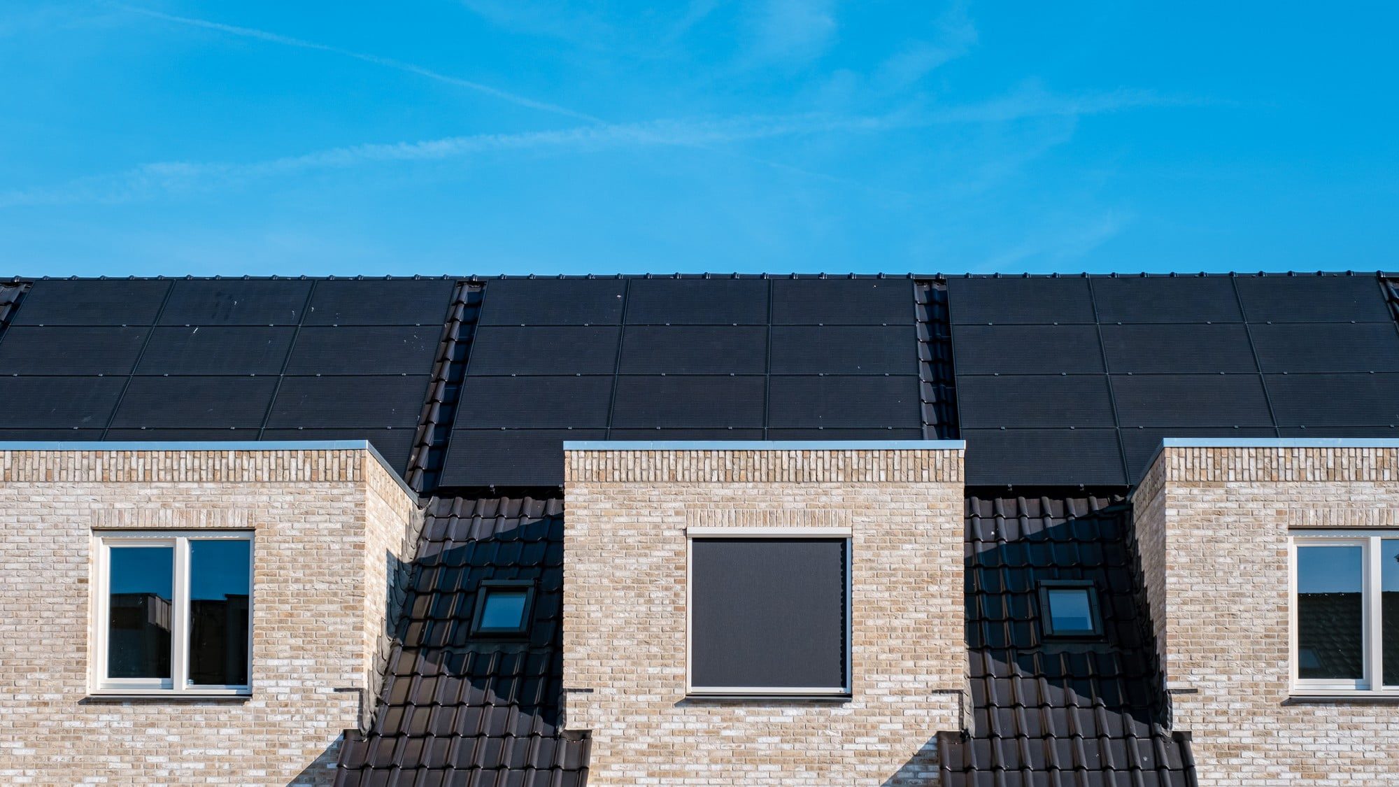 The image features a newly built residential area with rows of modern, detached houses. Each house has been outfitted with solar panels on its roof, highlighting a commitment to sustainable energy solutions. The houses are uniformly designed, giving a sense of cohesion and order to the neighborhood. The structures appear to be two-story buildings with a combination of light-coloured walls and darker roofing materials that contrast sharply with the glistening black surfaces of the solar panels. The roofing style is pitched, ideal for maximising solar energy capture.In the foreground, a well-maintained street curves gently, bordered by freshly planted grass and young trees, indicating that the area is newly developed. There are no cars or people visible, possibly suggesting the early morning or an off-peak time, contributing to the tranquil atmosphere of the scene. The sky is clear, hinting at favorable weather conditions, which enhances the overall brightness and clarity of the photograph. The image captures a sense of modern living with an emphasis on environmental consciousness.