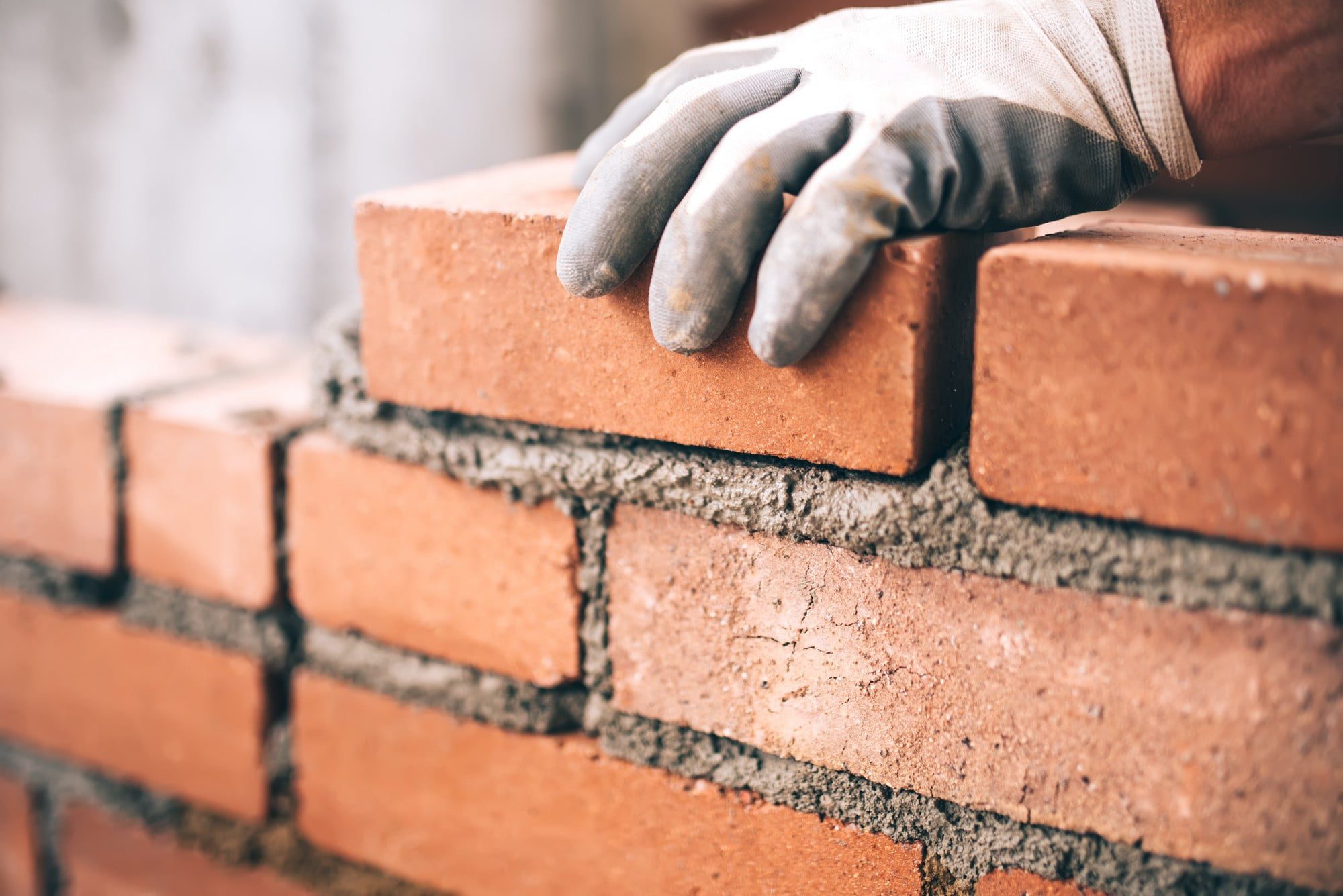 Alt text: "Close-up image of an industrial bricklayer installing a brick on a construction site. The bricklayer is wearing protective gloves and appears to be carefully placing a red brick onto a layer of wet mortar. The background shows a partially built brick wall, with other construction materials and tools visible, indicating active work. The bricklayer's focused expression and precise hand movements highlight the skill and attention to detail involved in the task."