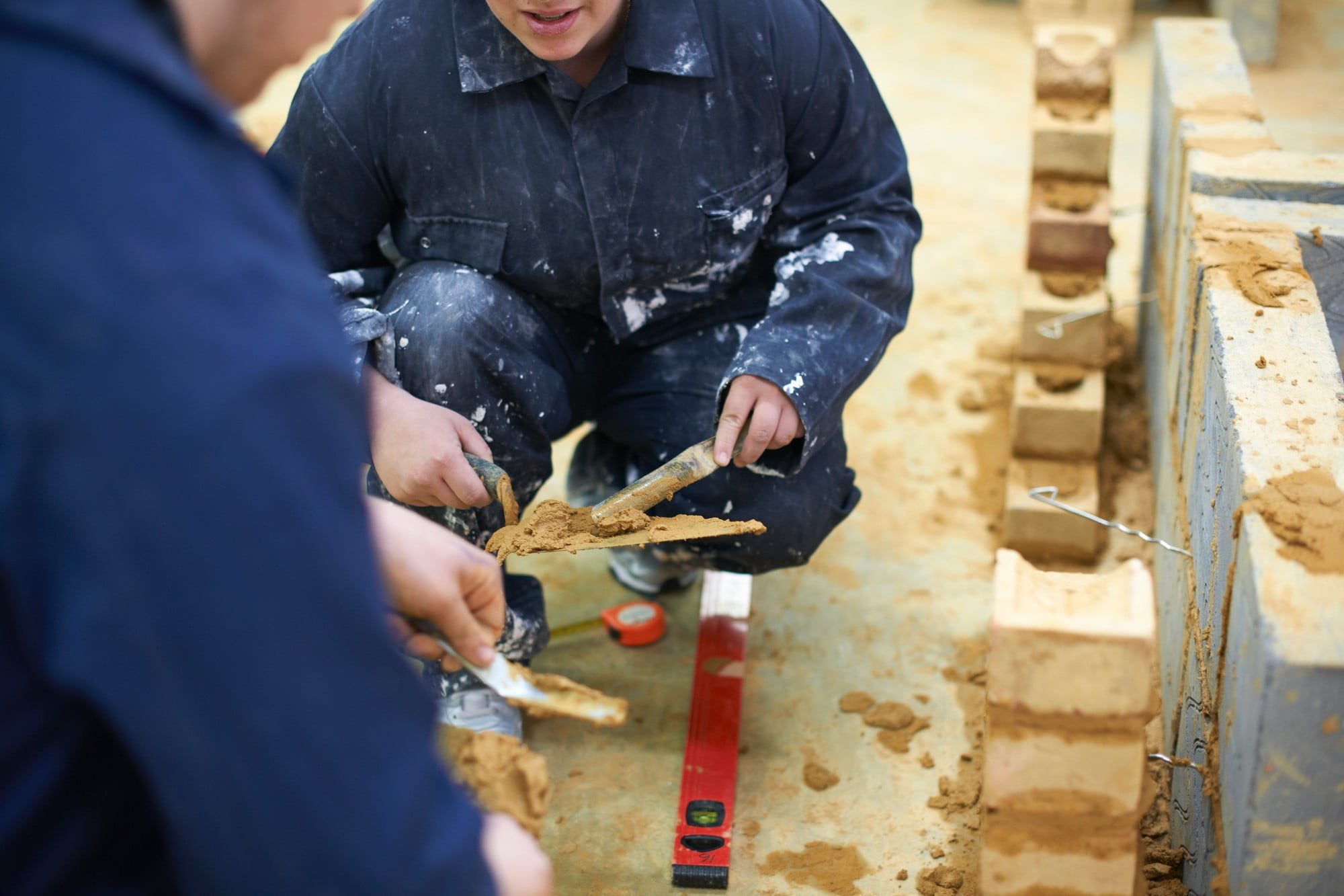 Alt text: A young student, wearing a yellow hard hat, safety goggles, and a high-visibility vest, is actively engaged in a hands-on building work lesson at a construction site. The student is using a trowel to apply mortar to a brick wall that is under construction. Other building materials and tools are visible around the site, including stacks of bricks and buckets. The background shows a partially completed structure, with scaffolding and some construction workers in the distance. The student appears to be concentrating on their task, demonstrating a focused and attentive demeanor. The scene is well-lit, suggesting it is taken during the daytime.